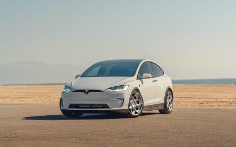 3/4 view of a light gray Tesla Model X featuring CYBRHEX Forged Wheels in Satin Titanium by Unplugged Performance, parked on a paved road with a vast desert landscape in the background under a clear blue sky.